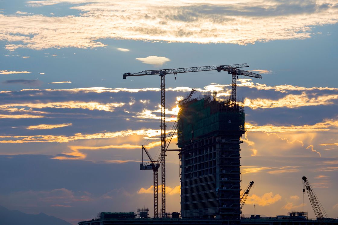 A high-rise building under construction, framed by two tower cranes against an evening sky.